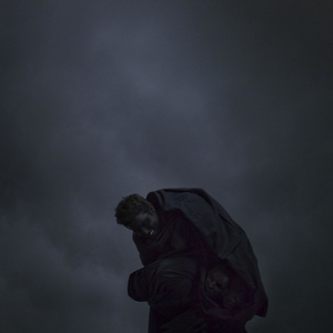 An upward shot of dark clouds. A person is wrapped in black cloth. Two other faces are visible peaking from within the folds. We can't see what these people are standing on, implying that it is a small towering platform.